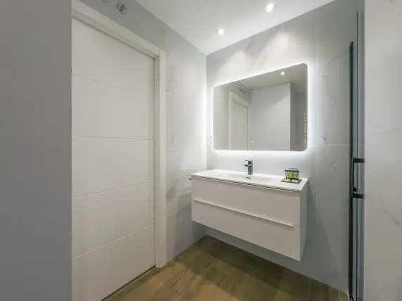 vista parcial del baño donde se observa el lavabo con mueble bajero y la zona de ducha de gran mampara acristalara que permite ver el interior de la ducha y el mármol blanco de veta gris de sus paredes elegante baño del apartamento de lujo Alfisa B1