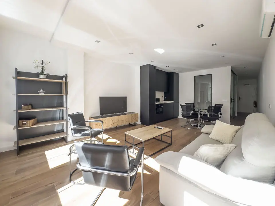 Vista panorámica del apartamento de lujo Alfisa desde el pasillo de entrada, mostrando la zona de estar con sofá, televisión y mueble enfrente. A la derecha, la mesa de comedor con sillas y la cocina en la pared. Al fondo, la zona de estar con mobiliario moderno, sofá cama grande y cómodo.