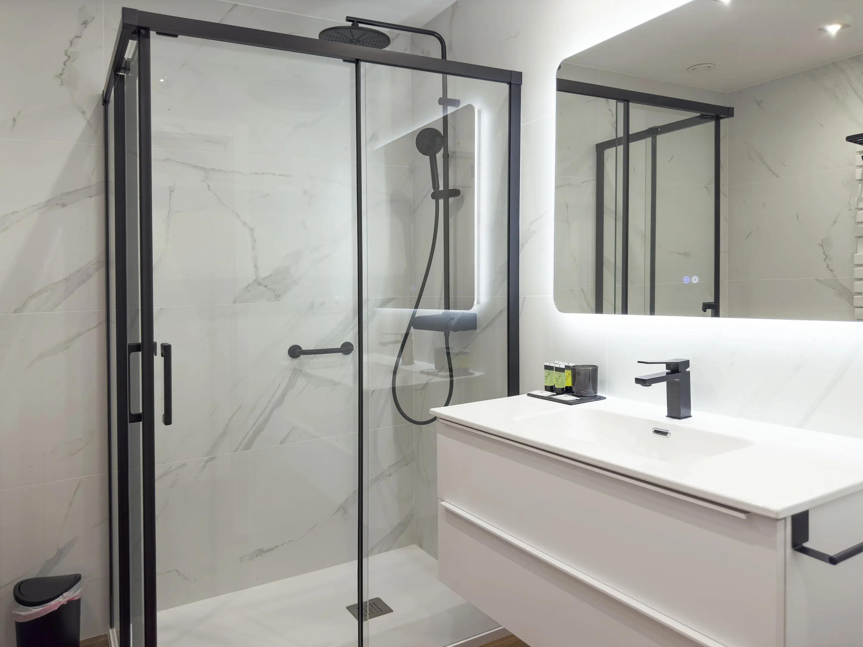 Vista parcial de un baño de lujo en los apartamentos Alfisa, con lavabo de mármol, grifo monomando, ducha con efecto lluvia y mamparas de cristal. Diseño moderno con espejo iluminado y detalles sofisticados