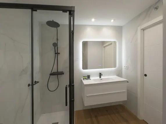 vista parcial donde aparece la ducha al fondo junto con el inodoro en la imagen se observa parte del lavabo con mueble bajero apartamento de lujo Alfisa B3