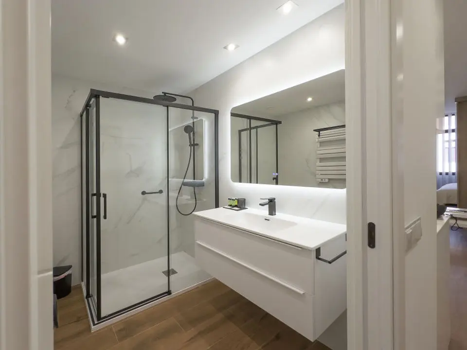 Vista parcial de un baño de lujo en los apartamentos Alfisa, con lavabo de mármol, grifo monomando, ducha con efecto lluvia y mamparas de cristal. Diseño moderno con espejo iluminado y detalles sofisticados