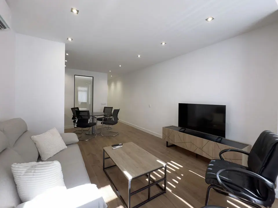Vista panorámica del apartamento de lujo Alfisa mostrando la zona de estar con sofá, televisión y mueble. A la izquierda la zona de estar con mobiliario moderno, sofá cama grande y cómodo. Al fondo, la mesa de comedor con sillas