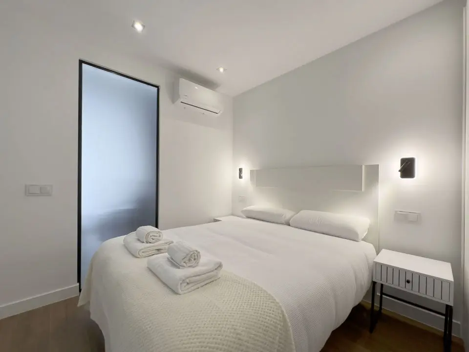 Habitación ultra moderna con cama de matrimonio en los apartamentos Alfisa, separada del resto de las instancias mediante una moderna estructura de madera, con mesillas a cada lado del cabecero , con luces individuales 