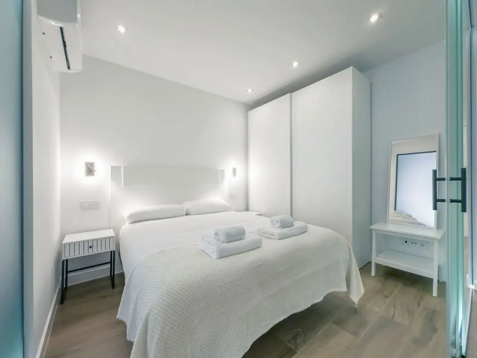 Habitación ultra moderna con cama de matrimonio en los apartamentos Alfisa, separada con grandes cristaleras traslúcidas del resto de las instancias, mesillas con luces individuales y armario de madera blanca al lado de la cama.