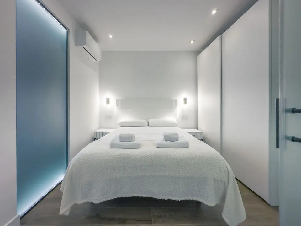 Habitación ultra moderna con cama de matrimonio en los apartamentos Alfisa, separada con grandes cristaleras traslúcidas del resto de las instancias, mesillas con luces individuales y armario de madera blanca al lado de la cama.