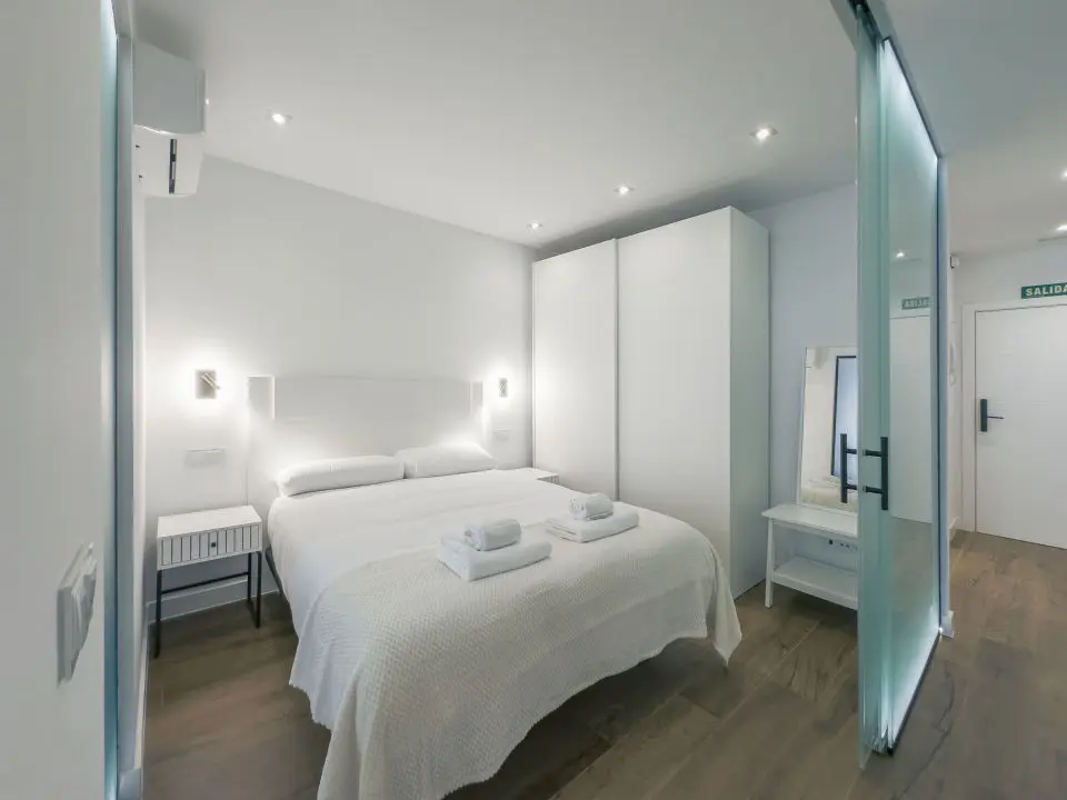 Habitación ultra moderna con cama de matrimonio en los apartamentos Alfisa, separada con grandes cristaleras traslúcidas del resto de las instancias, mesillas con luces individuales y armario de madera blanca al lado de la cama.