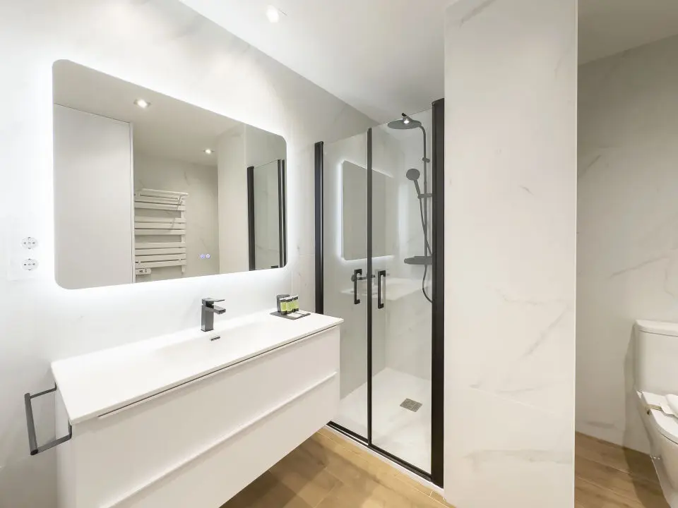 Vista parcial de un baño de lujo en los apartamentos Alfisa, con lavabo de mármol, grifo monomando, ducha con efecto lluvia y mamparas de cristal. Diseño moderno con espejo iluminado y detalles sofisticados