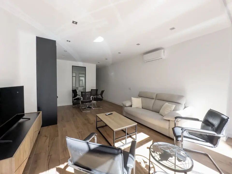 Vista panorámica del apartamento de lujo Alfisa desde el pasillo de entrada, mostrando la zona de estar con sofá, televisión y mueble enfrente. A la derecha, la mesa de comedor con sillas y la cocina en la pared. Al fondo, la zona de estar con mobiliario moderno, sofá cama grande y cómodo.