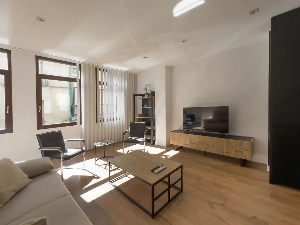 Vista panorámica del apartamento de lujo Alfisa desde el pasillo de entrada, mostrando la zona de estar con sofá, televisión y mueble enfrente. A la derecha, la mesa de comedor con sillas y la cocina en la pared. Al fondo, la zona de estar con mobiliario moderno, sofá cama grande y cómodo.