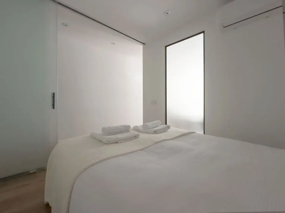 Habitación ultra moderna con cama de matrimonio en los apartamentos Alfisa, separada con grandes cristaleras traslúcidas del resto de las instancias, mesillas con luces individuales y armario de madera blanca al lado de la cama.