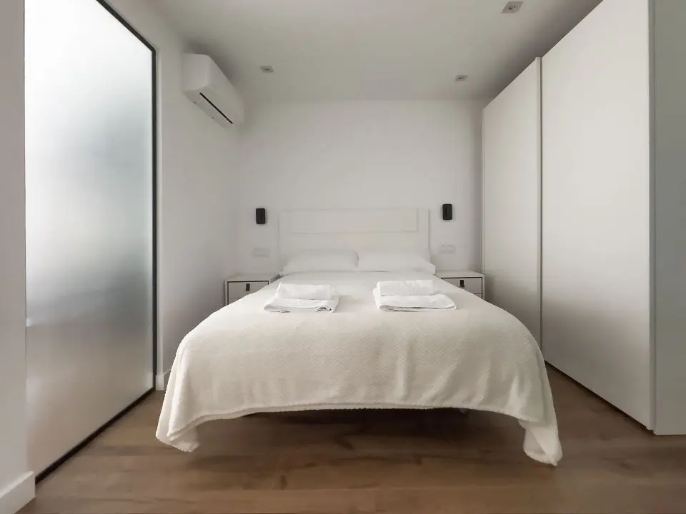 Habitación ultra moderna con cama de matrimonio en los apartamentos Alfisa, separada con grandes cristaleras traslúcidas del resto de las instancias, mesillas con luces individuales y armario de madera blanca al lado de la cama.