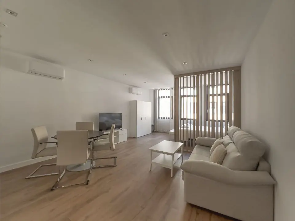 vista general del apartamento de lujo B3 de Alfisa, situado en l calle príncipe 5, a seis minutos a pie de la Puerta del Sol, se ve una cama con separación del resto de la estancia donde en un mismo espacio se sitúan el sofá cama, una mesa comedor con cuatro sillas, un mueble moderno bajo con televisor