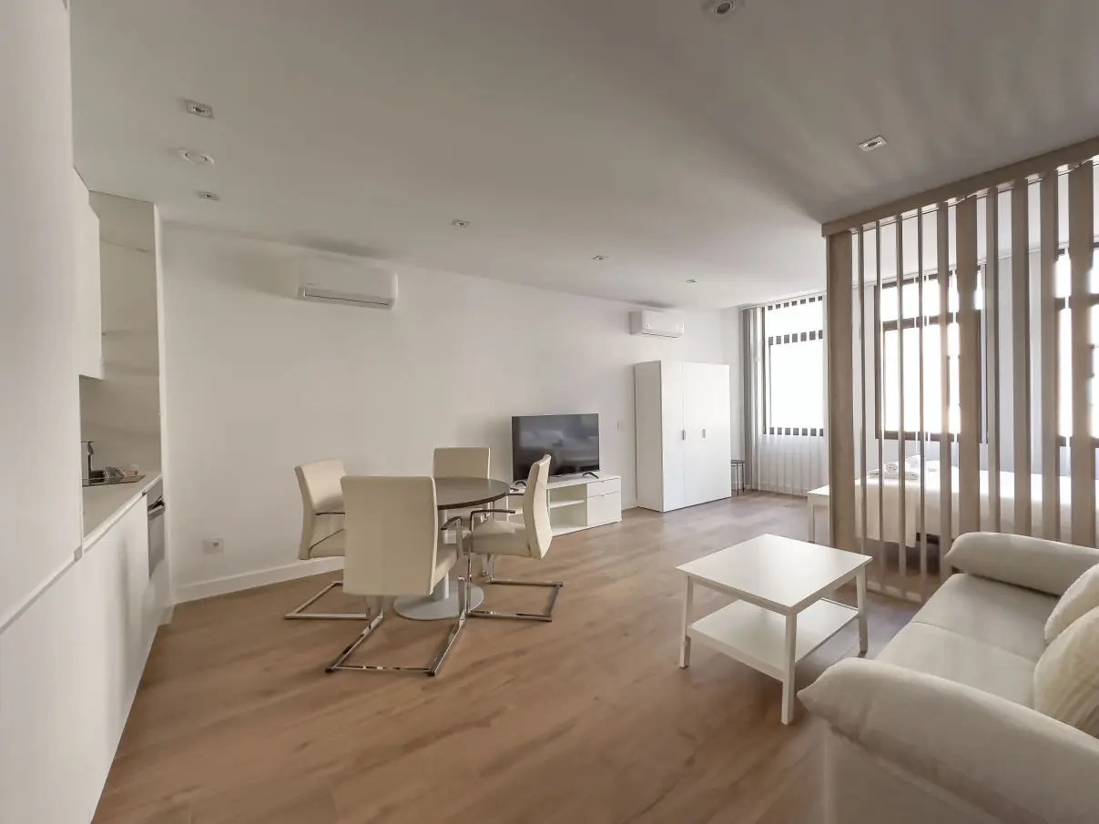 vista general del apartamento de lujo B3 de Alfisa, situado en l calle príncipe 5, a seis minutos a pie de la Puerta del Sol, se ve una cama con separación del resto de la estancia donde en un mismo espacio se sitúan el sofá cama, una mesa comedor con cuatro sillas, un mueble moderno bajo con televisor, y una cocina completa con nevera, horno, vitrocerámica y lavavajillas