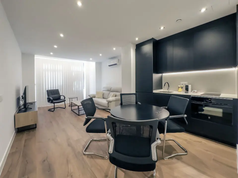 Vista panorámica del apartamento de lujo Alfisa desde el pasillo de entrada, mostrando la zona de estar con sofá cama, sillón de una plaza, mesa central pequeña y la televisión.