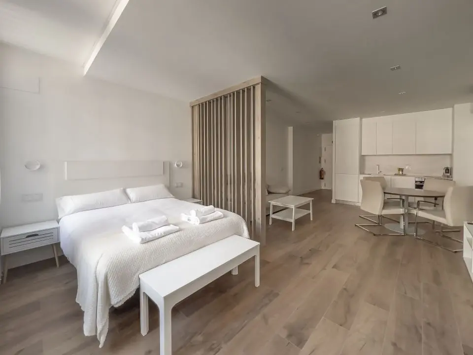 Vista panorámica del apartamento de lujo Alfisa desde la zona de habitación separada del resto de la estancia por una estructura  de madera que divide la cama de matrimonio. mostrando la zona de estar con sofá, televisión y mueble enfrente. A la derecha, la mesa de comedor con sillas y la cocina en la pared.