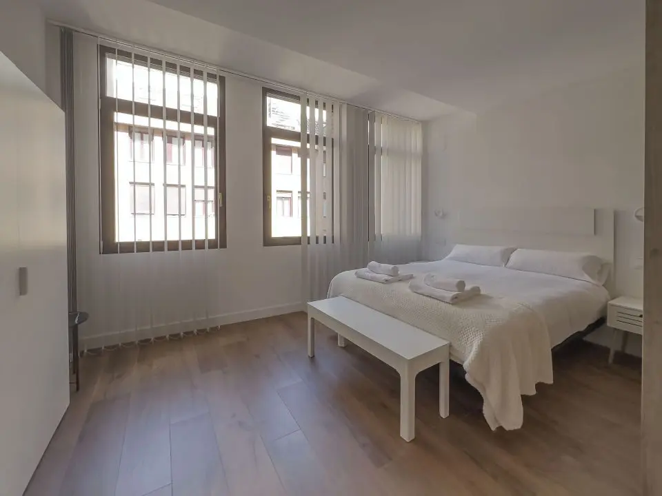 Habitación ultra moderna con cama de matrimonio en los apartamentos Alfisa, separada con grandes cristaleras traslúcidas del resto de las instancias, mesillas con luces individuales y armario de madera blanca al lado de la cama.