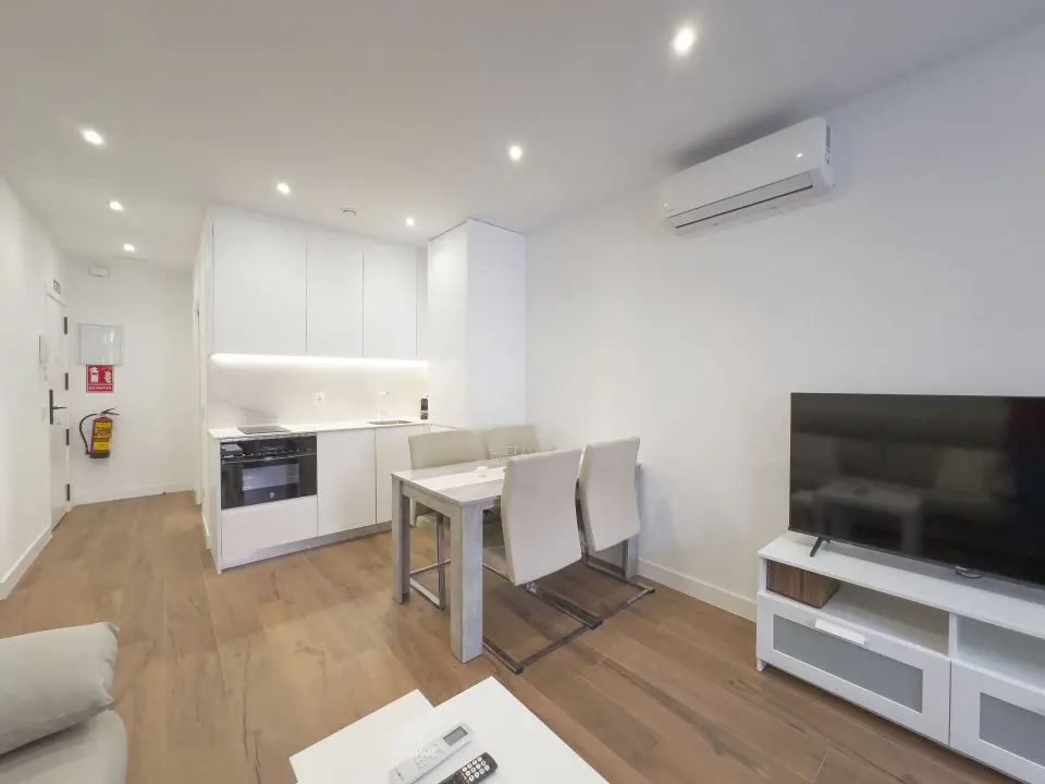 vista general del apartamento desde la habitación de matrimonio en los apartamentos Alfisa, grandes ventanales, mesillas con luces individuales y descalzador de madera blanca a los pies de la cama. Separador de madera artesanal en el lado derecho que divide la estancia del resto del apartamento.