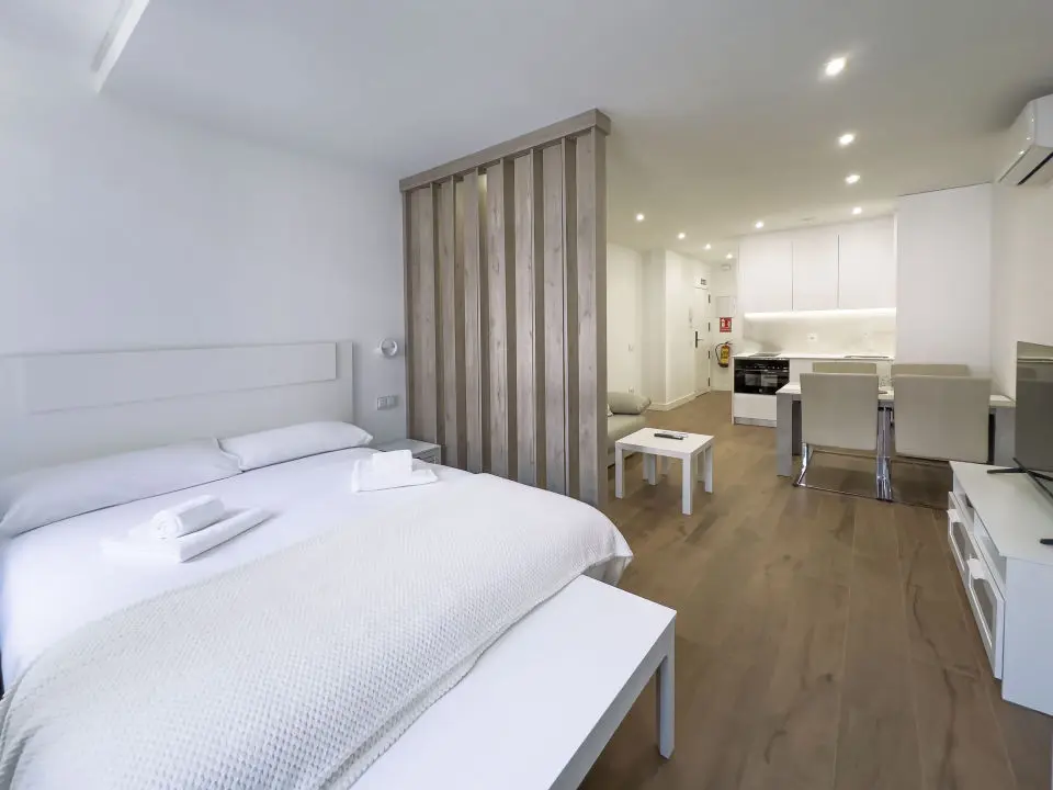 Habitación luminosa con cama de matrimonio en los apartamentos Alfisa, grandes ventanales, mesillas con luces individuales y descalzador de madera blanca a los pies de la cama. Separador de madera artesanal en el lado derecho que divide la estancia del resto del apartamento.