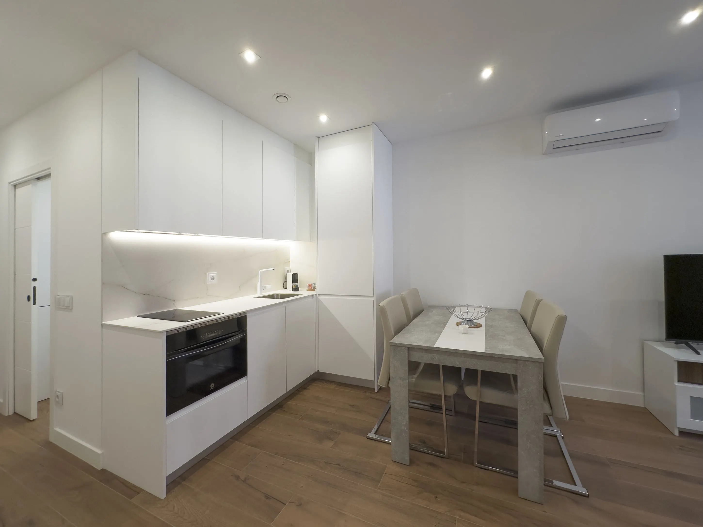 Cocina moderna en los apartamentos de lujo Alfisa, equipada con horno, vitrocerámica, cafetera y mesa de comedor con sillas. La nevera está integrada y oculta. También se aprecia el sistema de aire acondicionado, en un espacio con diseño contemporáneo.