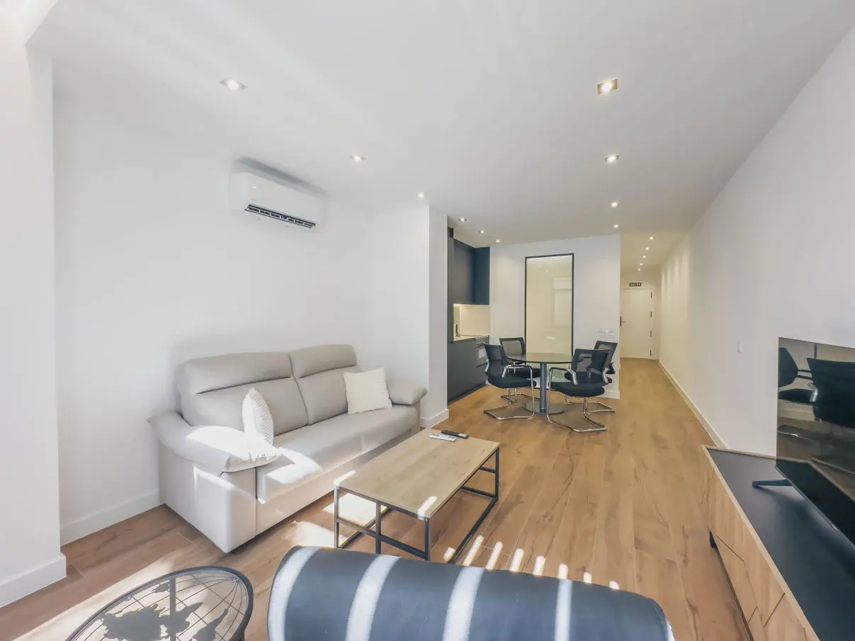 vista general del apartamento de lujo B1 de Alfisa, situado en l calle príncipe 5, a seis minutos a pie de la Puerta del Sol, se ve una cama con separación del resto de la estancia donde en un mismo espacio se sitúan el sofá cama, una mesa comedor con cuatro sillas, un mueble moderno bajo con televisor, y una cocina completa con nevera, horno, vitrocerámica y lavavajillas, habitación con grandes cristaleras